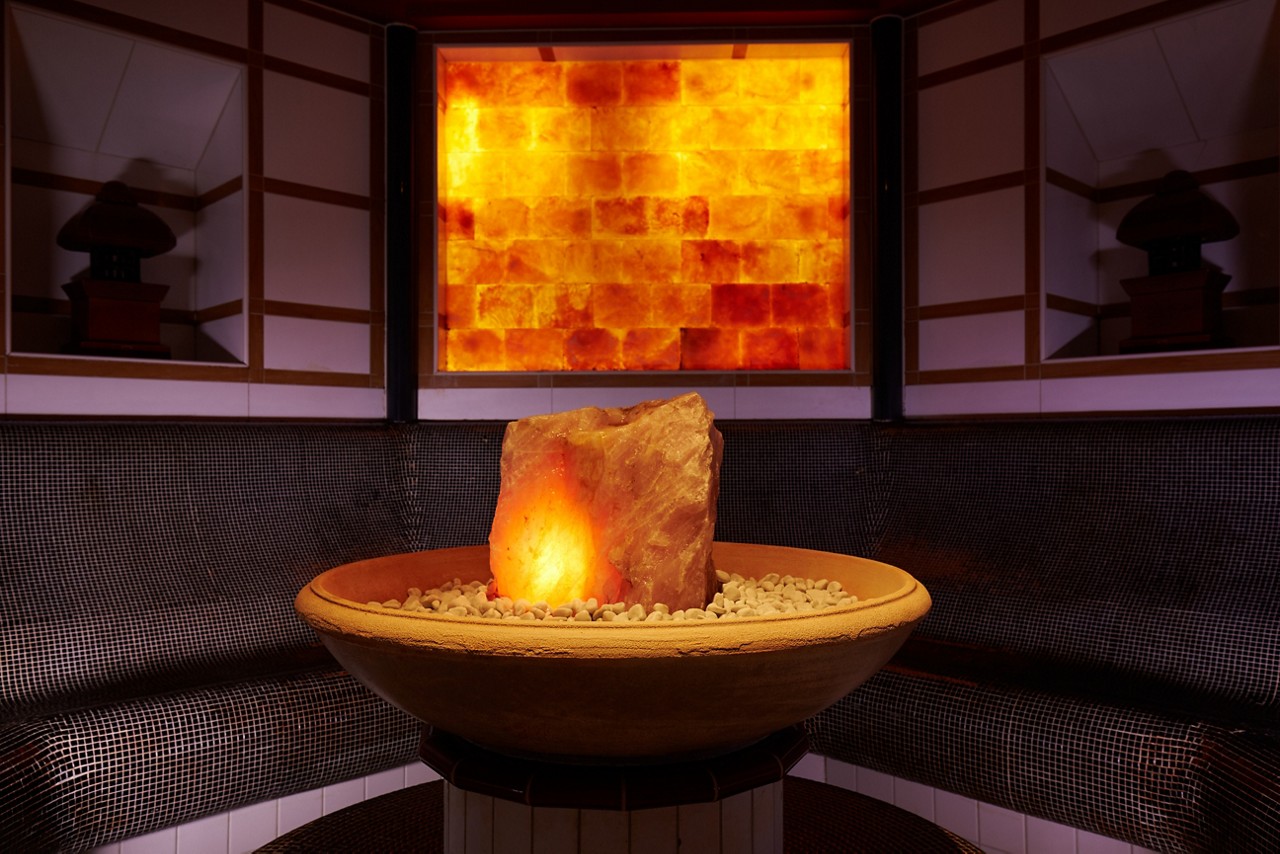 A glowing salt rock sits in a pebble-filled bowl, illuminating a dark tiled steam room with curved bench seating and a backlit amber brick wall.
